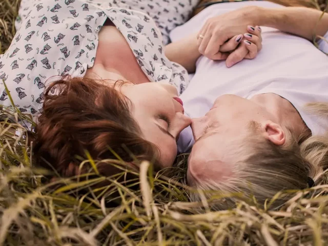 Ein Paar liegt mit geschlossenen Augen auf einer Wiese, während seine Hände und Nasenspitzen sich berühren.