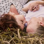 Ein Paar liegt mit geschlossenen Augen auf einer Wiese, während seine Hände und Nasenspitzen sich berühren.