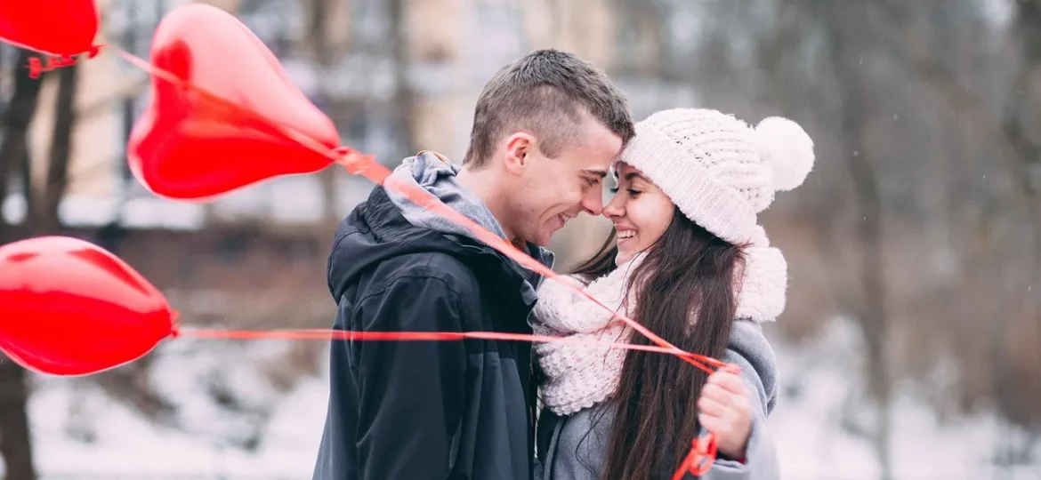 Die erste der 5 Phasen einer Beziehung: Ein Paar hält verliebt die Nasenspitzen aneinander, während sie drei Herzballons in der Hand hält.