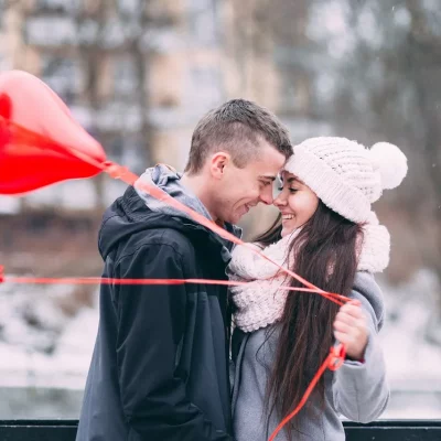 Die erste der 5 Phasen einer Beziehung: Ein Paar hält verliebt die Nasenspitzen aneinander, während sie drei Herzballons in der Hand hält.