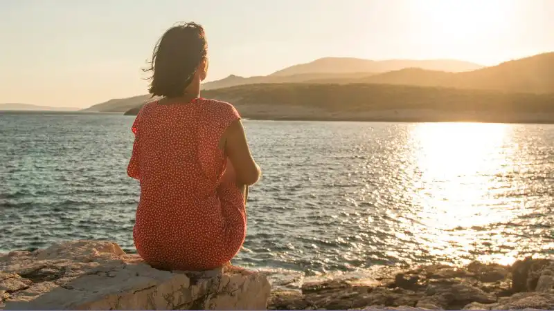 Person sitzt allein am Wasser bei Sonnenuntergang – Symbol für Selbstreflexion und Mitgefühl im Kurs Liebe ohne Kampf